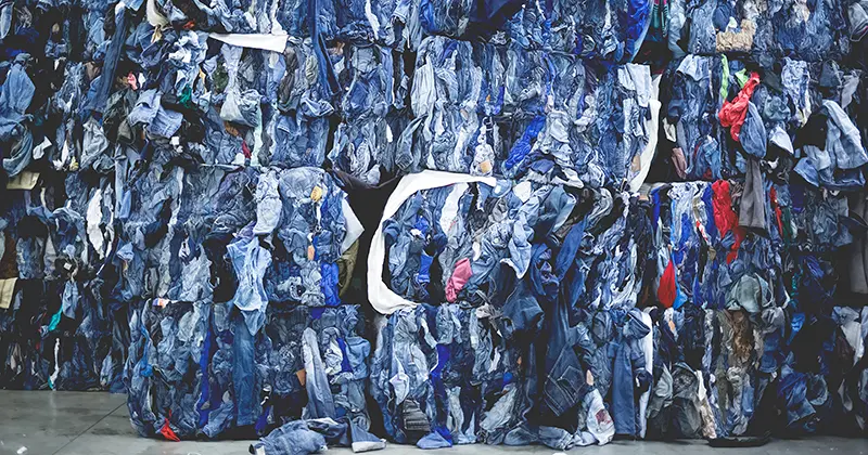 In een magazijn liggen verschillende grote balen met stevig samengeperste, gerecyclede blauwe denimstof en kleding opgestapeld.